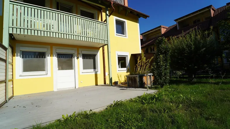 Gelbes Reihenhaus mit Balkon und einer gepflasterten Terrasse im Erdgeschoss, umgeben von Grünflächen.