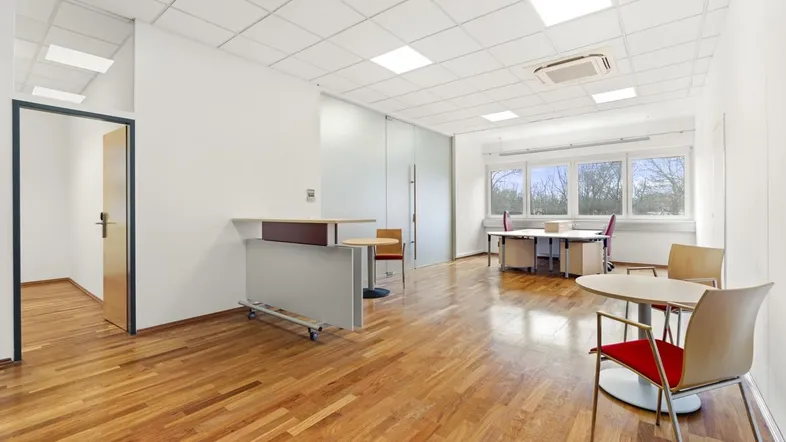 Geräumiges Büro mit hellem Holzfußboden, großen Fenstern und moderner Deckenbeleuchtung.