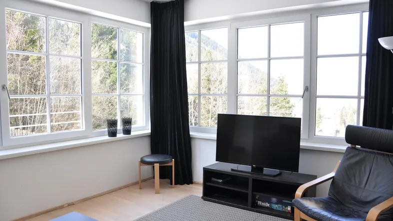 Helles Wohnzimmer mit großen Fenstern, Bergblick und dunklen Vorhängen.