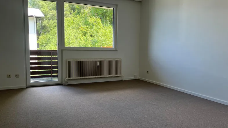Helles Zimmer mit Teppichboden, weiß gestrichenen Wänden und Zugang zu einem Balkon mit Blick ins Grüne.
