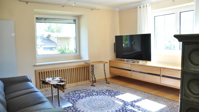 Helles Wohnzimmer mit Holzboden, großem Teppich, modernem TV-Möbel und Kachelofen.
