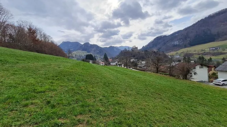 Weitläufige grüne Wiese mit Blick auf ein malerisches Dorf und die umliegende Berglandschaft unter bewölktem Himmel.
