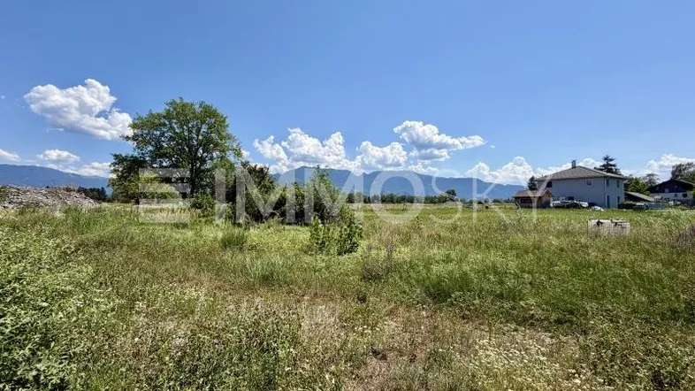 Weitläufiges Grundstück mit grüner Wiese, umgeben von Bergen und vereinzelten Häusern unter blauem Himmel.