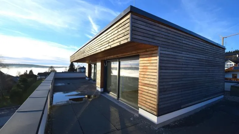 Moderne Architektur mit Holzfassade und großer Dachterrasse unter blauem Himmel.