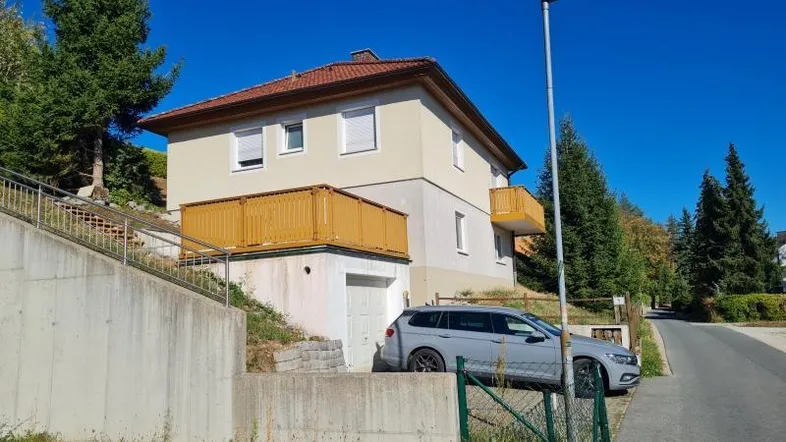 Einfamilienhaus mit beiger Fassade, rotem Ziegeldach und angrenzender Straße an einem sonnigen Tag.