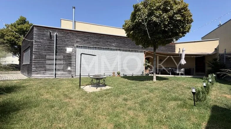 Moderner Gartenbereich mit Holzfassade, Rasenfläche, Grillplatz und überdachter Terrasse.