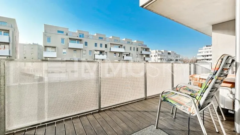 Geräumiger Balkon mit Holzboden und Blick auf moderne Wohngebäude unter blauem Himmel.