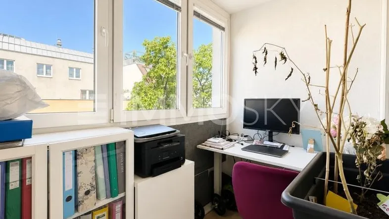 Helles Büro mit Schreibtisch, Drucker und Blick ins Grüne durch große Fenster.