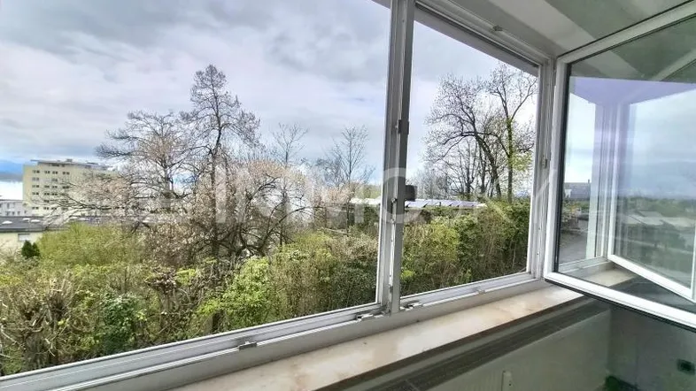 Blick aus dem Fenster eines Wohnraums mit grüner Umgebung und fernen Gebäuden.