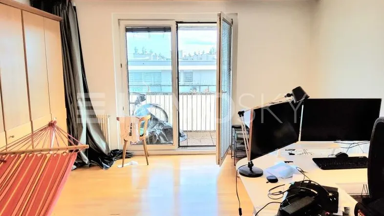 Helles Zimmer mit Holzboden, Schreibtisch und Zugang zu einem kleinen Balkon mit Stadtblick.