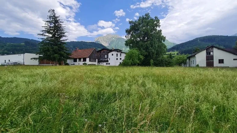 Weitläufiges grünes Feld mit Blick auf umliegende Häuser und majestätische Berge unter blauem Himmel.