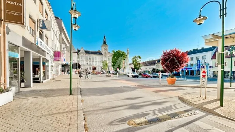 Belebter Stadtplatz mit historischen Gebäuden, Geschäften und einem Uhrturm unter blauem Himmel.