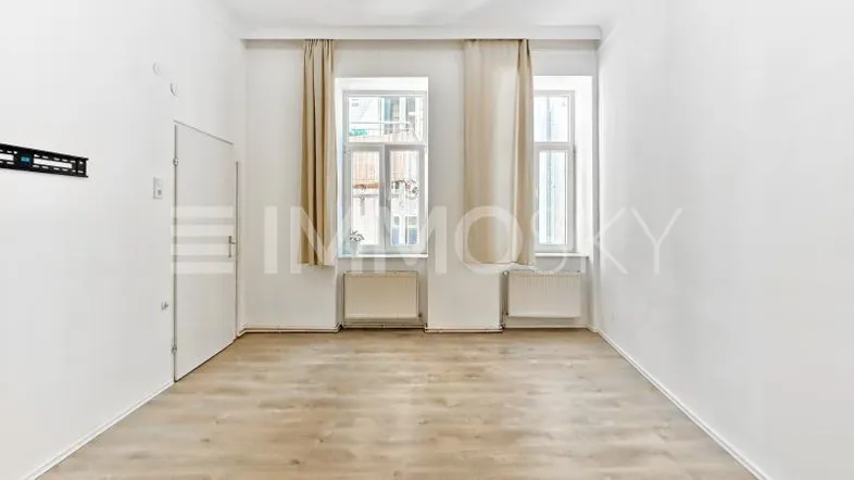 Helles, leeres Zimmer mit Laminatboden, weißen Wänden und zwei Fenstern mit Vorhängen.