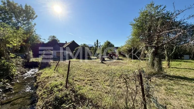 Sonniges Grundstück mit Bachlauf und Blick auf die umliegende Landschaft und ein Gebäude im Hintergrund.