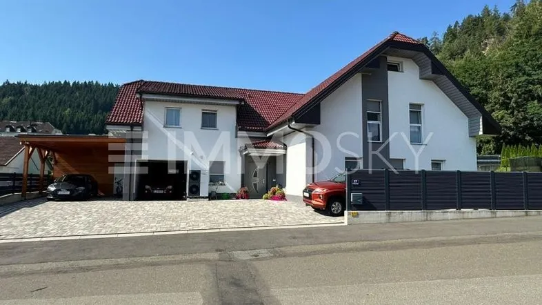 Modernes Einfamilienhaus mit Satteldach, Carport und gepflasterter Auffahrt in grüner Umgebung.