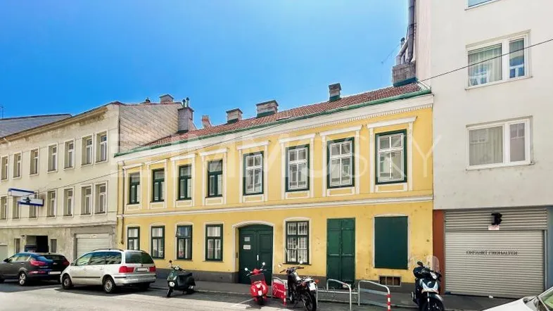Historisches Gebäude mit gelber Fassade, grünen Fensterläden und parkenden Fahrzeugen an einer städtischen Straße.