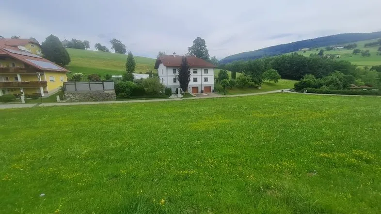 Weitläufige grüne Wiese mit Blick auf ländliche Gebäude und sanfte Hügel in der Ferne.