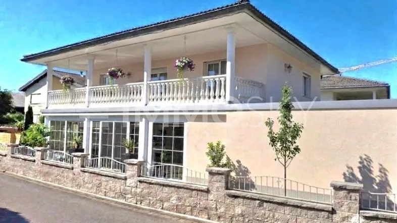 Elegantes Einfamilienhaus mit großem Balkon, Säulen und Steinmauer im Vorgartenbereich.