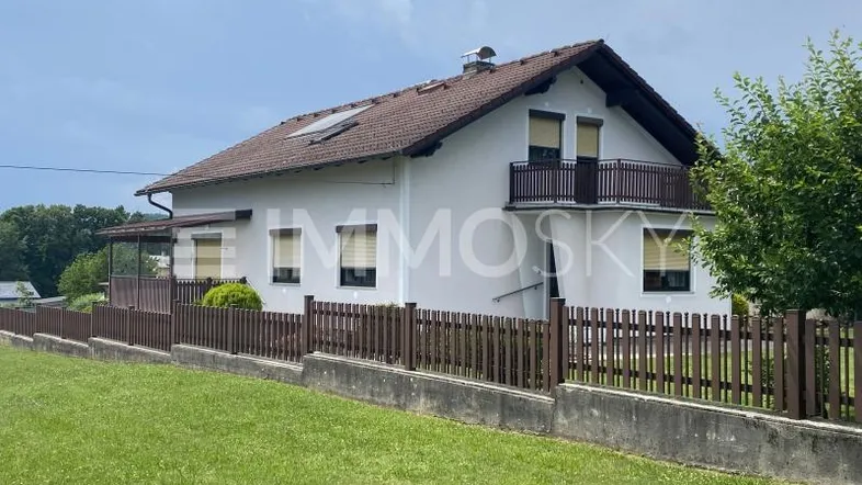Einfamilienhaus mit Satteldach, Balkon und umzäuntem Vorgarten in ländlicher Umgebung.