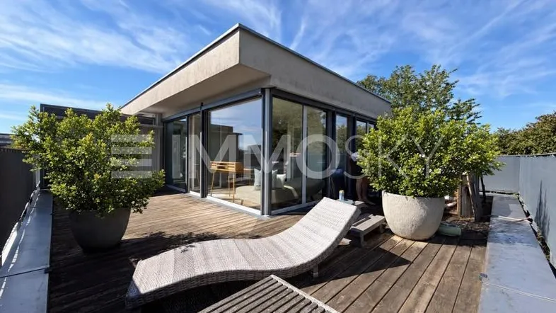 Geräumige Dachterrasse mit Holzdeck, Liegestuhl und modernen Glasfronten des Gebäudes unter blauem Himmel.