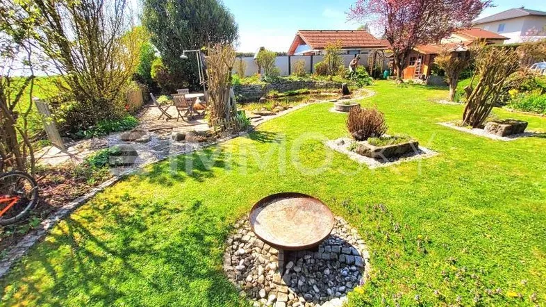 Großer, gepflegter Garten mit Rasenfläche, Teich, Feuerstelle und Sitzbereich unter blauem Himmel.