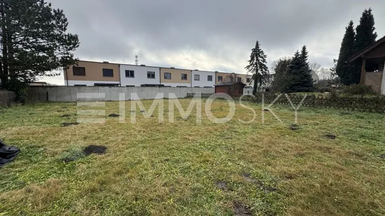 Ungenutztes Grundstück mit Grasfläche und Blick auf moderne Reihenhäuser unter bewölktem Himmel.
