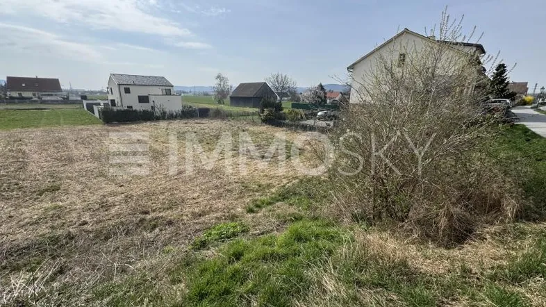 Unbebautes Grundstück mit trockenem Gras, umgeben von Wohnhäusern und ländlicher Umgebung.