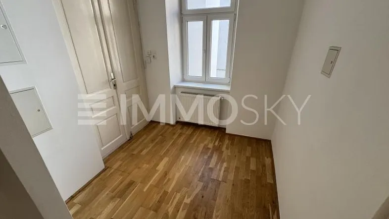 Helles Zimmer mit Parkettboden, weiß gestrichenen Wänden und einem Fenster, das Tageslicht hereinlässt.