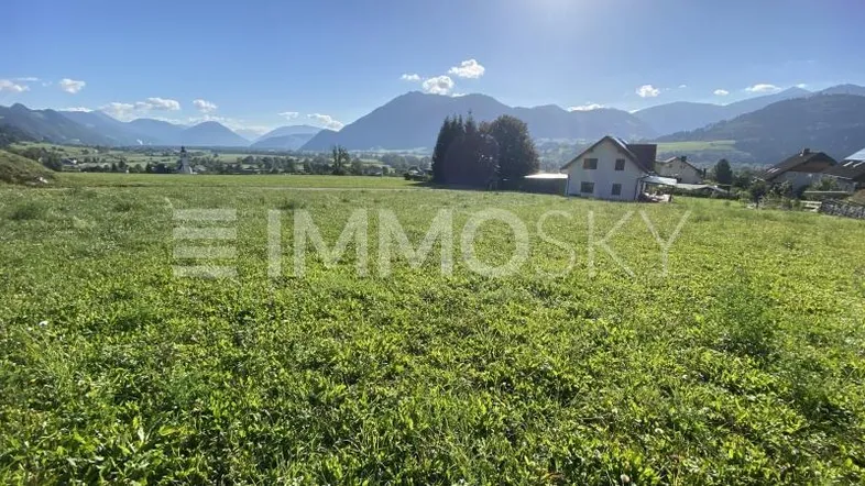 Weitläufige grüne Landschaft mit Bergblick und einem Haus im Hintergrund unter blauem Himmel.