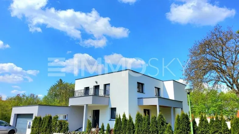 Modernes weißes Einfamilienhaus mit Garage, kleinem Balkon und gepflegter Vorgartenbepflanzung unter blauem Himmel.