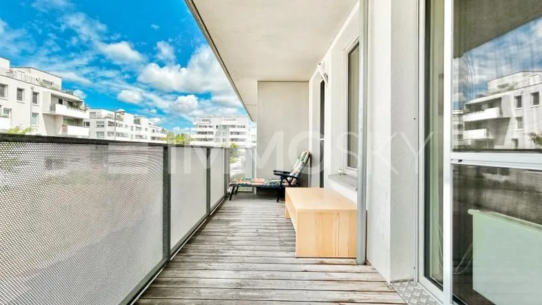 Langer Balkon mit Holzboden, Sitzgelegenheiten und Blick auf die umliegenden Gebäude.