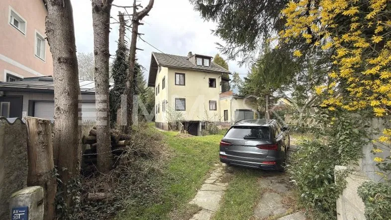 Einfamilienhaus mit Zufahrt und Stellplatz, umgeben von Bäumen und Sträuchern in ländlicher Umgebung.