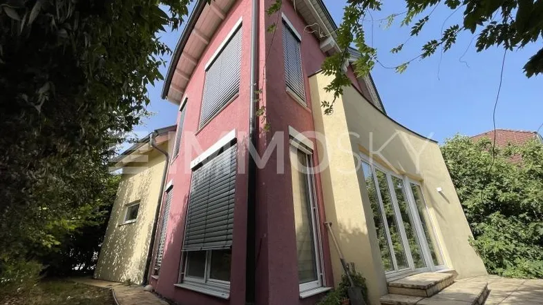 Außenansicht eines Hauses mit markanter rosa und gelber Fassade, umgeben von üppigem Grün.