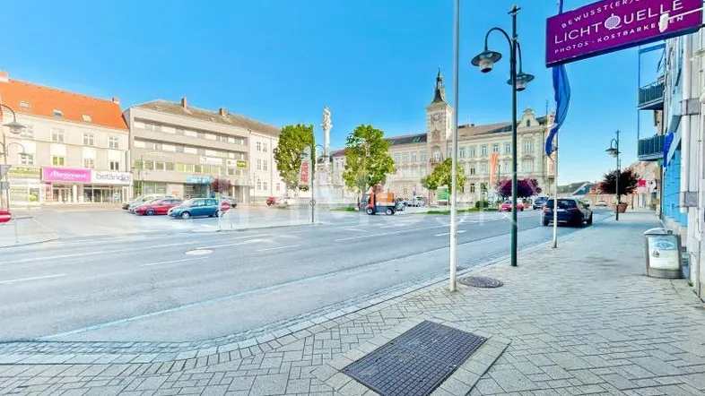 Stadtplatz mit historischen Gebäuden, gepflasterten Gehwegen und blauem Himmel an einem sonnigen Tag.