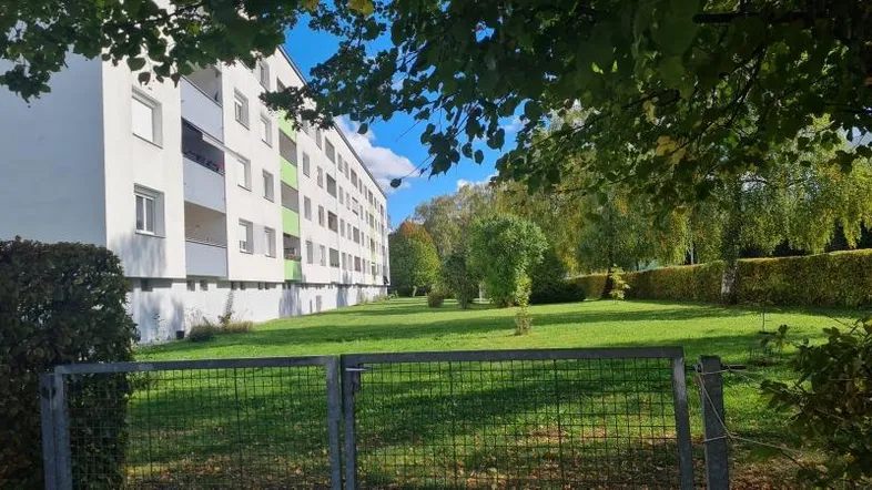 Mehrfamilienhaus mit großer Grünfläche und Bäumen, umgeben von einem Zaun.