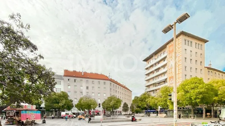 Blick auf einen belebten Stadtplatz mit umliegenden Gebäuden, Bäumen und einem Fahrradständer.