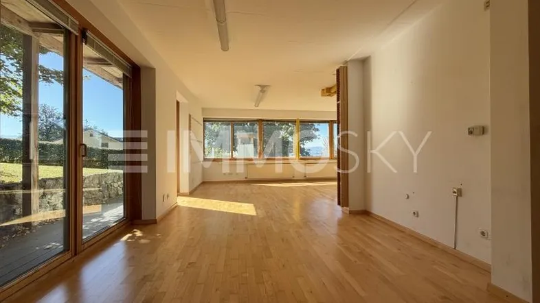 Helles, leeres Wohnzimmer mit Holzboden und großen Fenstern, die viel Tageslicht hereinlassen.