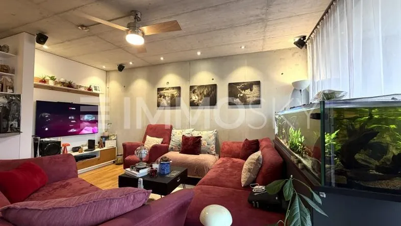 Geräumiges Wohnzimmer mit bequemen Sofas, Holzboden, Betondecke und einem großen Aquarium.