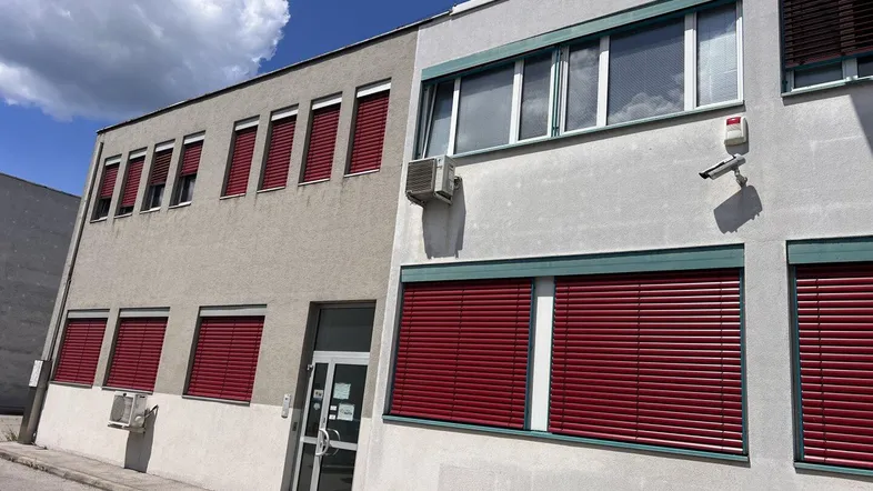 Gebäudeaußenansicht mit mehreren Fenstern, roten Jalousien und einer Überwachungskamera an der Fassade.