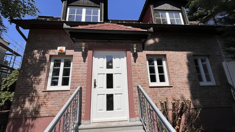 Vorderansicht eines traditionellen Backsteinhauses mit weißer Eingangstür und Dachgauben unter blauem Himmel.