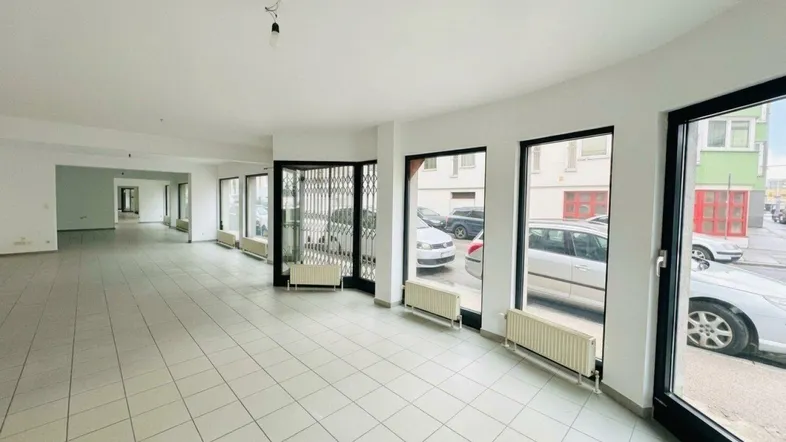 Geräumiger Gewerberaum mit großen Schaufenstern, Fliesenboden und Heizkörpern, ideal für Einzelhandel oder Büro.