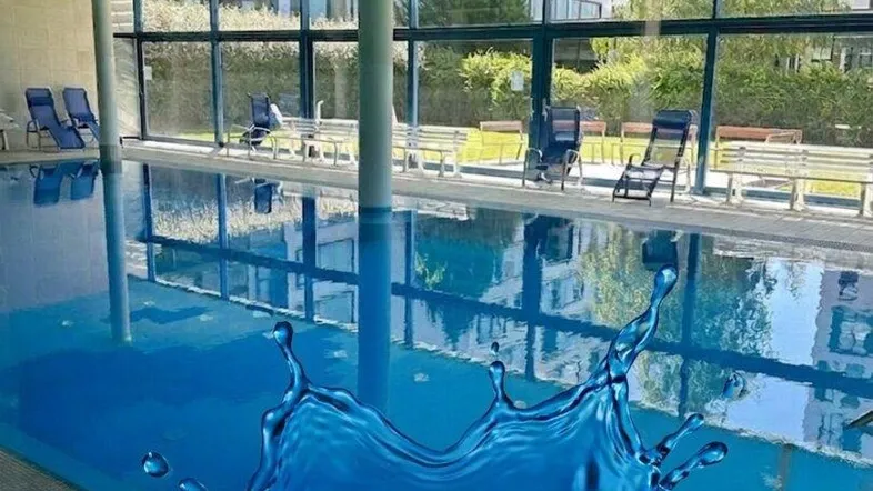 Helles Hallenbad mit großen Fenstern, Liegestühlen und Blick ins Grüne, Teil einer Wellnessanlage.