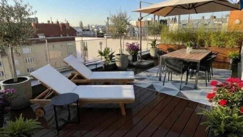 Geräumige Dachterrasse mit Holzdeck, Sonnenliegen, Esstisch und weitem Stadtblick unter blauem Himmel.