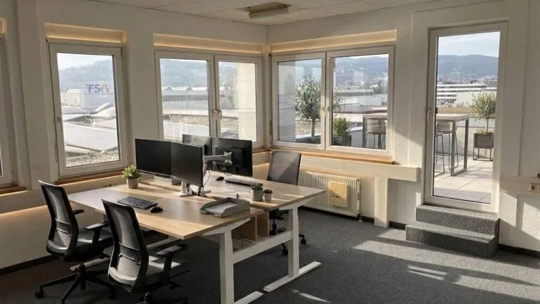 Modernes Büro mit großen Fenstern, viel Tageslicht und weitem Stadt- und Bergblick. Ergonomische Arbeitsplätze und direkter Zugang zu einer Dachterrasse.