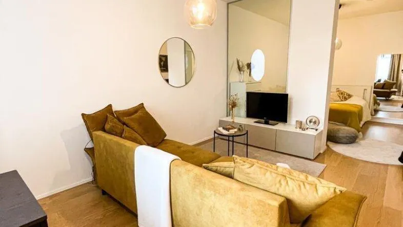 Helles Wohnzimmer mit modernem Sofa, Holzboden und offenem Übergang zum Schlafbereich.