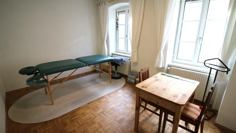 Helles Therapiezimmer mit Massageliege, Holztisch und zwei Fenstern, ideal für Behandlungen oder Beratungen.