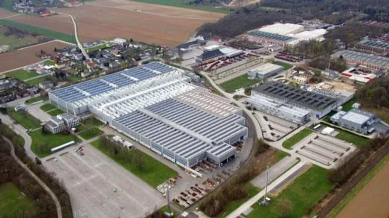 Luftaufnahme eines großen Industriegebiets mit mehreren Hallen und Photovoltaikanlagen auf den Dächern, umgeben von Feldern und Wald.