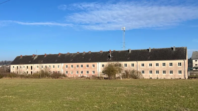 Langes Wohngebäude mit vielen Fenstern und einem weiten Feld im Vordergrund unter blauem Himmel.