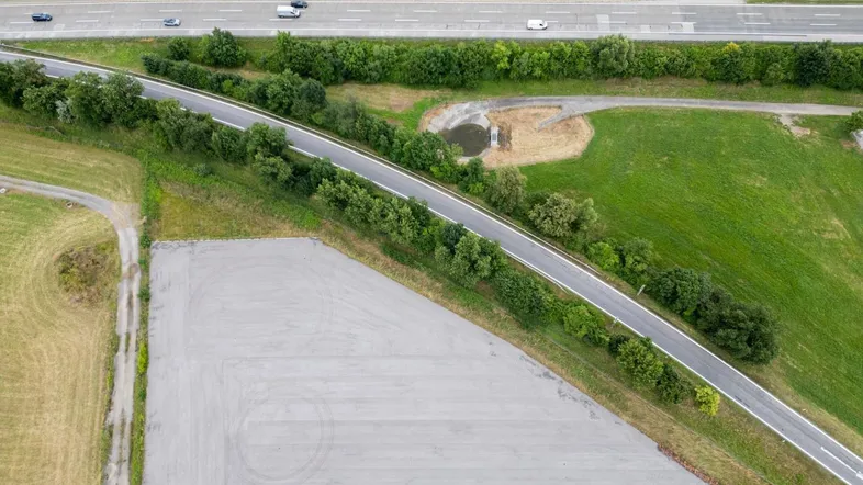 Luftaufnahme eines großen asphaltierten Bereichs neben einer Autobahn, umgeben von grünen Feldern und Bäumen.