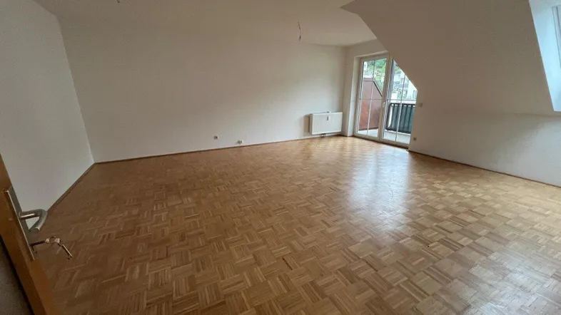 Helles Zimmer mit Parkettboden, Dachschräge und Zugang zu einem kleinen Balkon.
